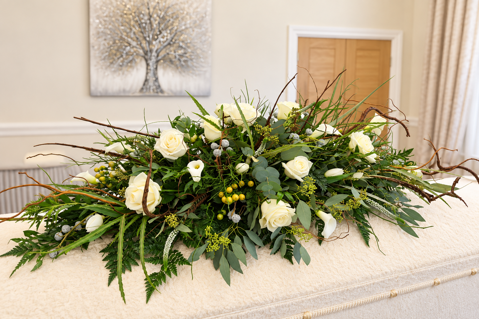 White roses and green foliage resting on a white coffin in a quiet room