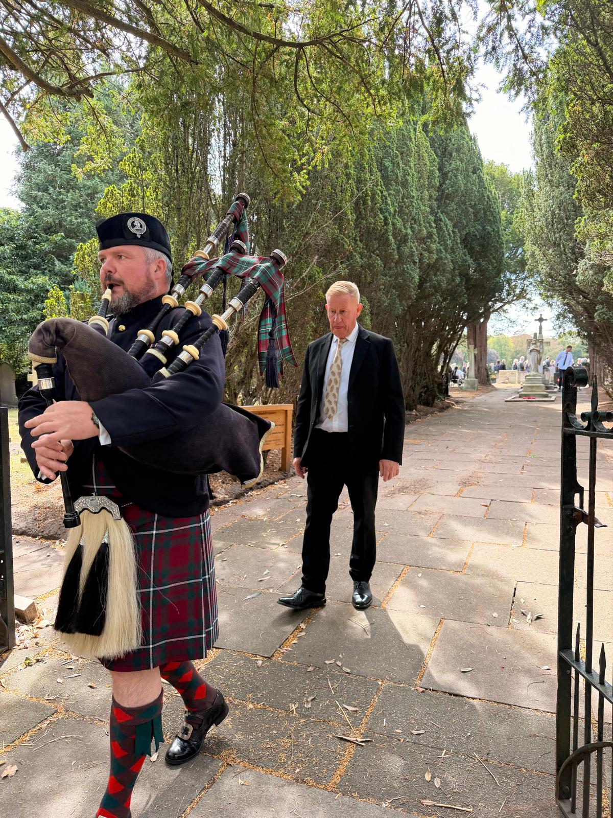 A bagpiper leading a funeral procession beneath trees